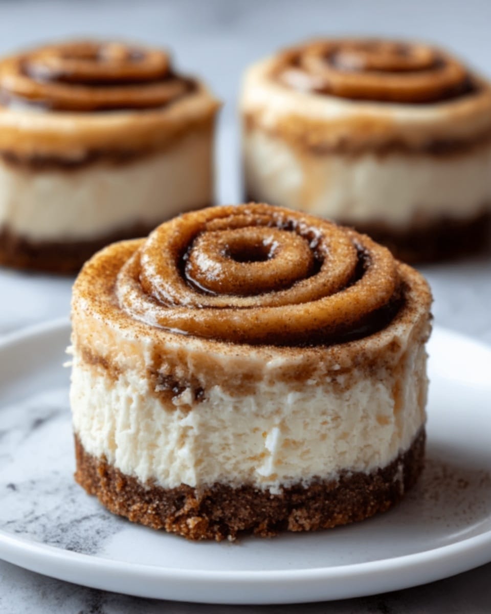 The image shows a close-up of a small cheesecake with three visible layers on a white marbled surface. The bottom layer is a dark brown crumbly crust, the middle layer is a smooth, creamy light beige cheesecake, and the top layer is a tightly swirled cinnamon roll with a golden brown color and visible cinnamon texture. There is a thick layer of white cream between the cheesecake and the cinnamon roll, with some cream lightly spilling over the edge. The background includes two more cheesecakes slightly out of focus. Photo taken with an iphone --ar 4:5 --v 7