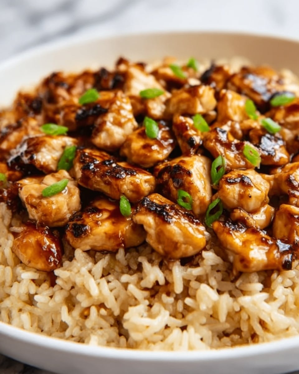 The image shows a close-up of a dish with two main layers. The bottom layer is fluffy white rice with a slight shine, filling the whole frame at the base. On top, there are chunks of grilled chicken, cooked to a golden brown with dark grill marks, arranged closely together. The chicken pieces are coated with a glossy, dark sauce that makes them look juicy and rich. Small green garnishes are scattered lightly over the chicken, adding a fresh color contrast. The background has a white marbled texture. Photo taken with an iphone --ar 4:5 --v 7