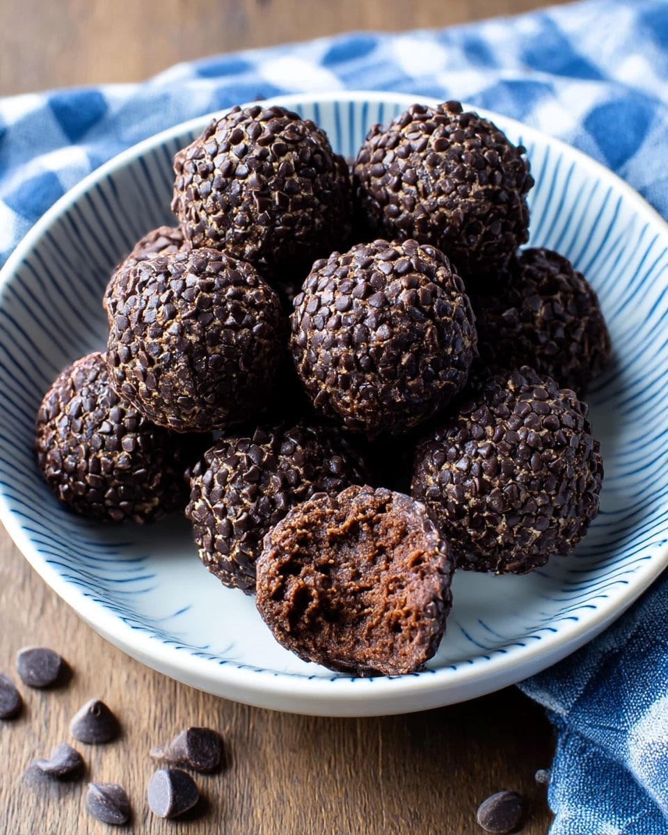 A white bowl with thin blue stripes holds about ten round chocolate balls covered fully in small, dark chocolate chips; one ball is slightly bitten, showing a dense, dark brown inside that looks soft and moist. The balls have a rough, textured surface due to the chips, and a few chocolate chips are scattered around the bowl on a wooden surface, which is partially covered by a blue and white checkered cloth. photo taken with an iphone --ar 4:5 --v 7