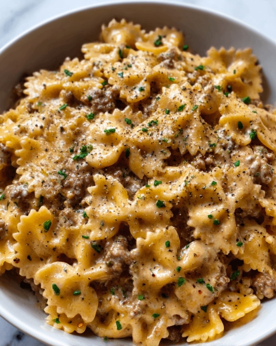 The image shows a close-up of a white bowl filled with pasta shaped like small bow ties, mixed with ground meat. The pasta is coated in a creamy light brown sauce, and small bits of chopped green herbs are sprinkled evenly on top. Some black pepper is scattered over the dish, adding texture and contrast. The pasta pieces look tender and slightly glossy, while the ground meat appears browned and crumbly. The bowl is set on a white marbled surface, and the photo focuses closely on the food, showing the creamy sauce clinging to the pasta and meat. Photo taken with an iphone --ar 4:5 --v 7