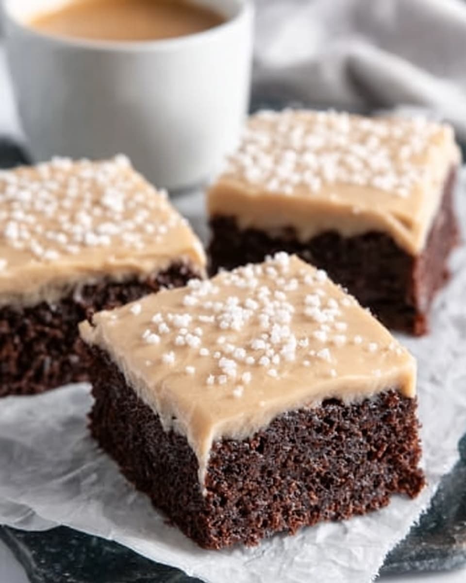 The image shows three square chocolate brownies with a thick layer of light caramel-colored frosting on top, sprinkled with small white sugar crystals. The brownies have a dark, rich brown base that looks moist and dense. The frosting layer is smooth and glossy, covering each brownie fully, and the sugar crystals add a bit of texture and sparkle. They are placed on white parchment paper, and in the background, there's a blurred white cup filled with a light brown drink, all set against a white marbled surface. photo taken with an iphone --ar 4:5 --v 7