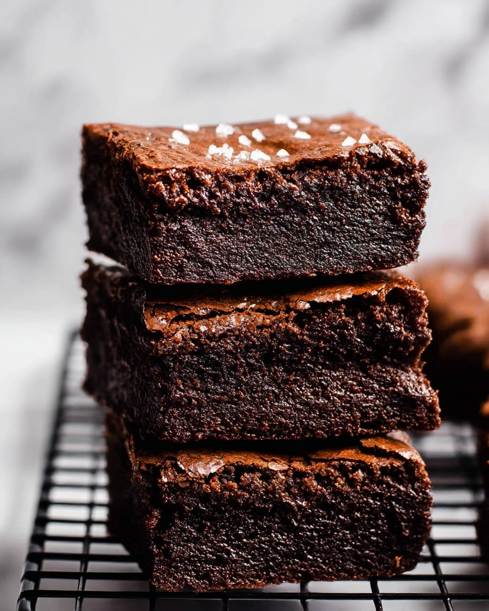 The image shows four thick rectangular chocolate brownies stacked neatly on top of each other, each with a dark, rich color and a slightly cracked top crust with a shiny texture. The sides reveal a dense, moist interior with a deep brown color. The brownies are placed on a black wire cooling rack, set against a white marbled surface in the background. Small white sugar crystals are lightly sprinkled on top, adding contrast to the dark chocolate. Photo taken with an iphone --ar 4:5 --v 7