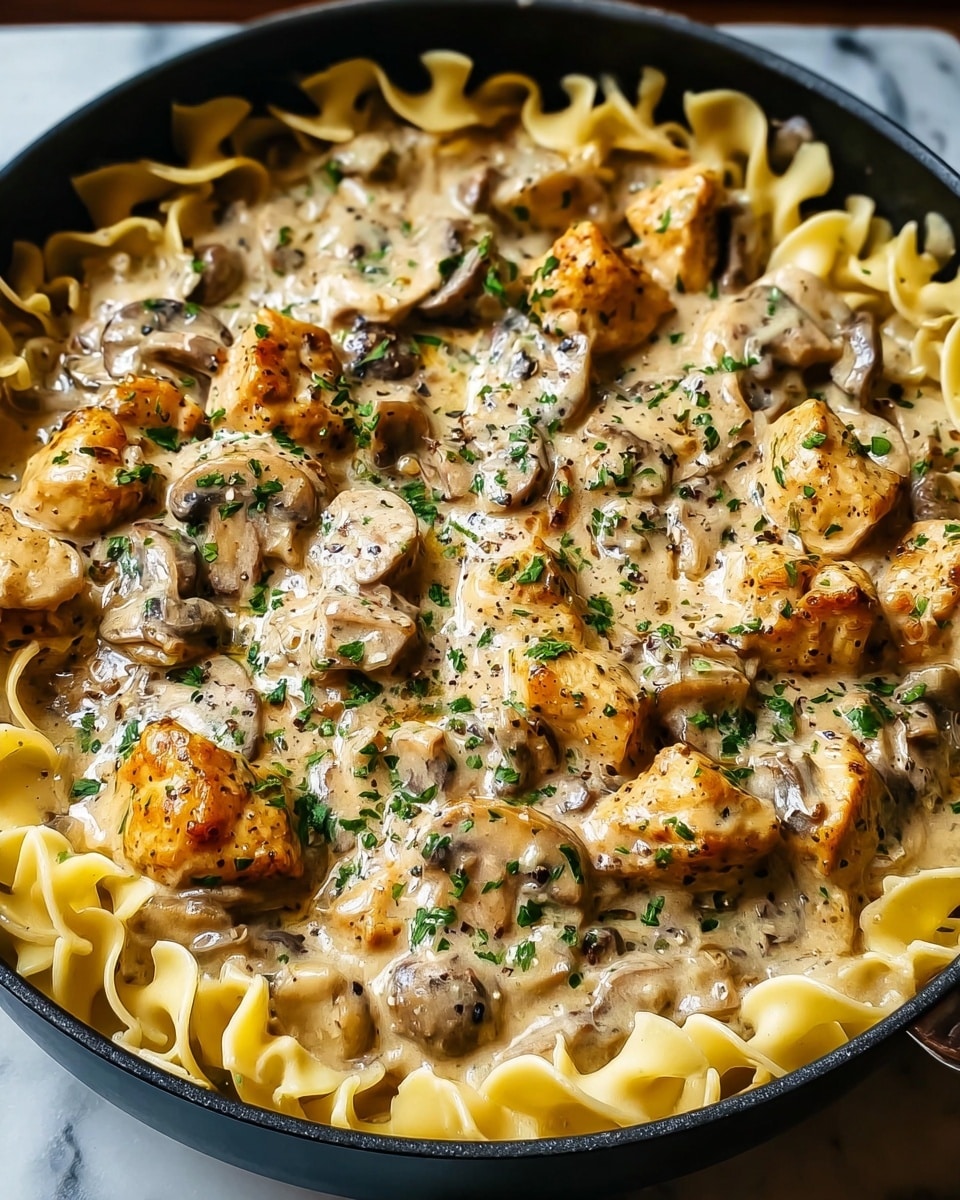 The dish shows a close-up of a black pan filled with three layers: the bottom layer is light beige egg noodles with a wavy texture, the middle layer is a creamy sauce with visible small black pepper flakes and herbs, and the top layer has browned chicken chunks and sliced mushrooms coated in the sauce, garnished with finely chopped green herbs scattered evenly across the surface, all set on a white marbled textured background. photo taken with an iphone --ar 4:5 --v 7
