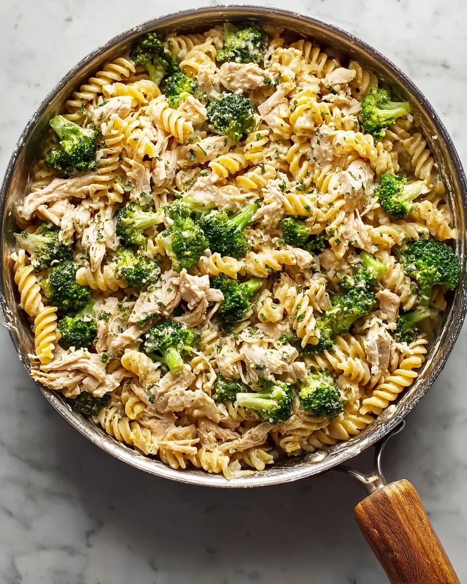 A large round pan filled with a creamy pasta dish that has three main layers visible: the first layer is yellow spiral rotini pasta, the second layer is bright green broccoli florets scattered evenly, and the third layer is shredded pieces of light brown cooked chicken mixed throughout. The dish looks well-mixed with small bits of herbs sprinkled on top, and the pan handle is wooden, resting on a white marbled surface. The textures show creamy sauce coating the pasta and chicken, giving a slightly glossy finish. photo taken with an iphone --ar 4:5 --v 7