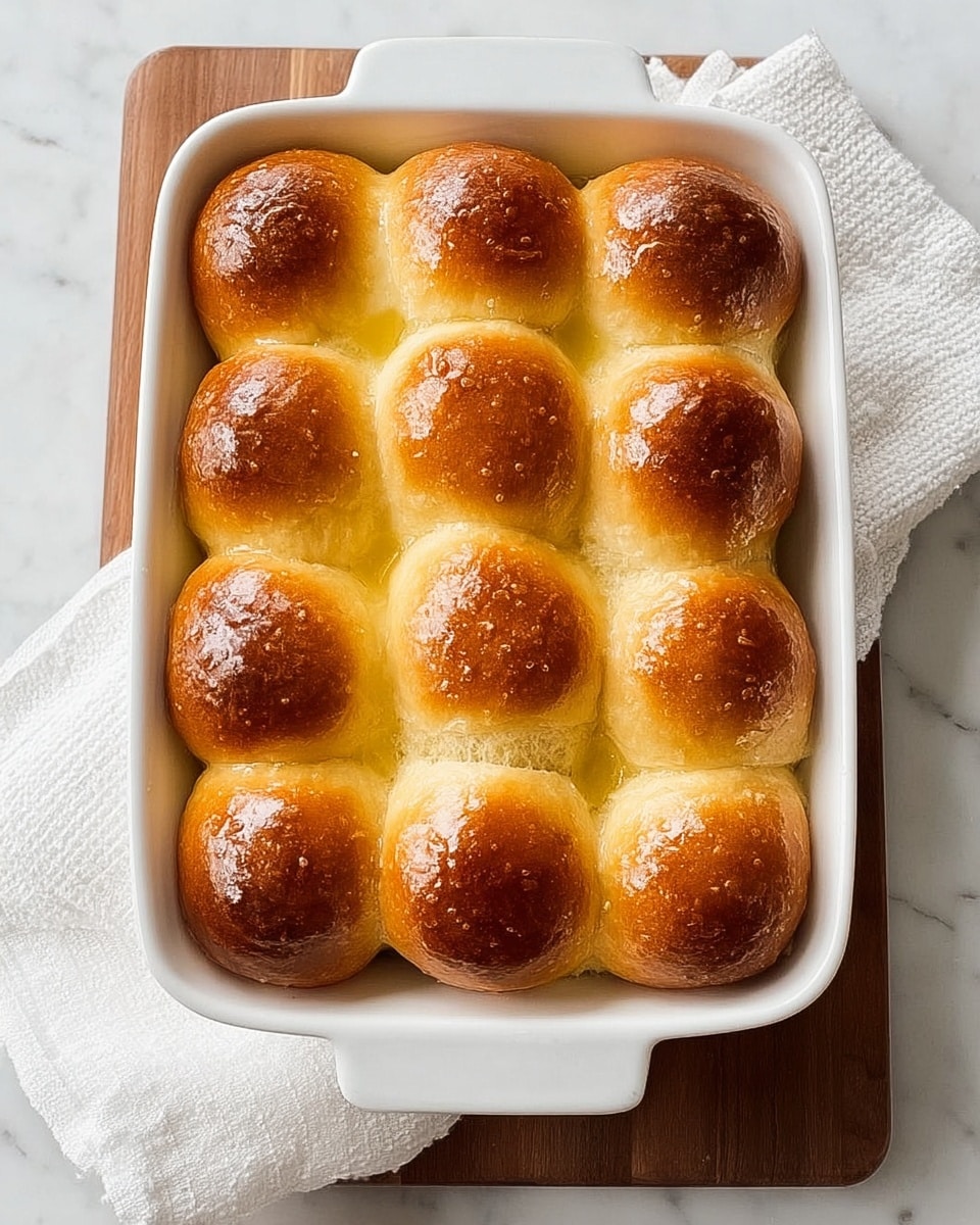 A white rectangular baking dish holding twelve soft, round bread rolls arranged in a 3 by 4 grid; each roll has a shiny, golden-brown top with a slightly darker hue around the edges and a fluffy, light yellow base visible between them; the dish sits on a white textured cloth placed on a white marbled surface with a wooden board underneath. photo taken with an iphone --ar 4:5 --v 7