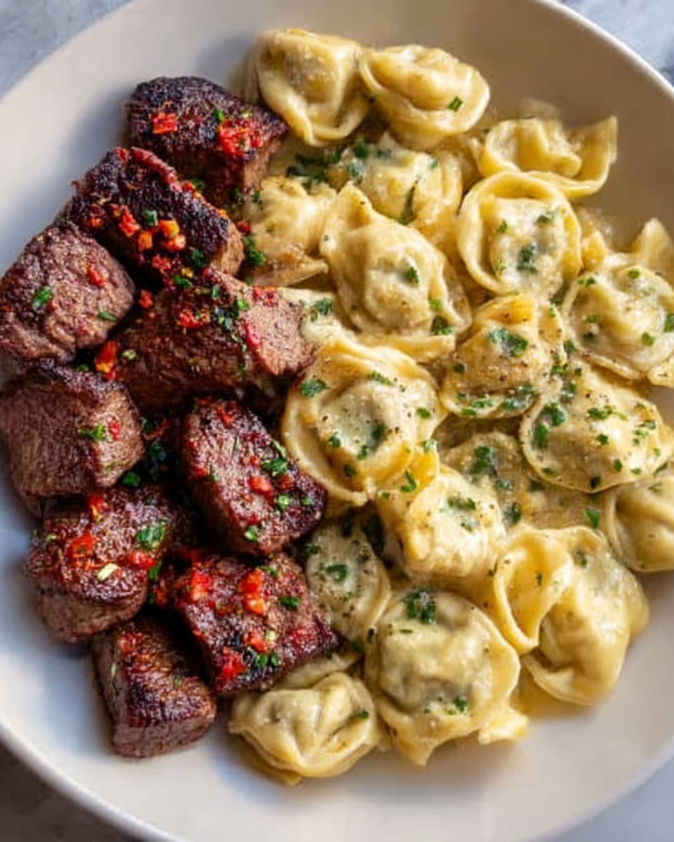 The image shows a white plate filled with two main sections: on one side, there are several pieces of grilled steak cubes with a slightly charred surface and reddish-brown color, seasoned with herbs and small red pepper bits. On the other side, there is a generous serving of cheese tortellini pasta, light yellow in color with a smooth, creamy sauce and garnished with chopped green herbs. The plate rests on a white marbled surface. Photo taken with an iphone --ar 4:5 --v 7