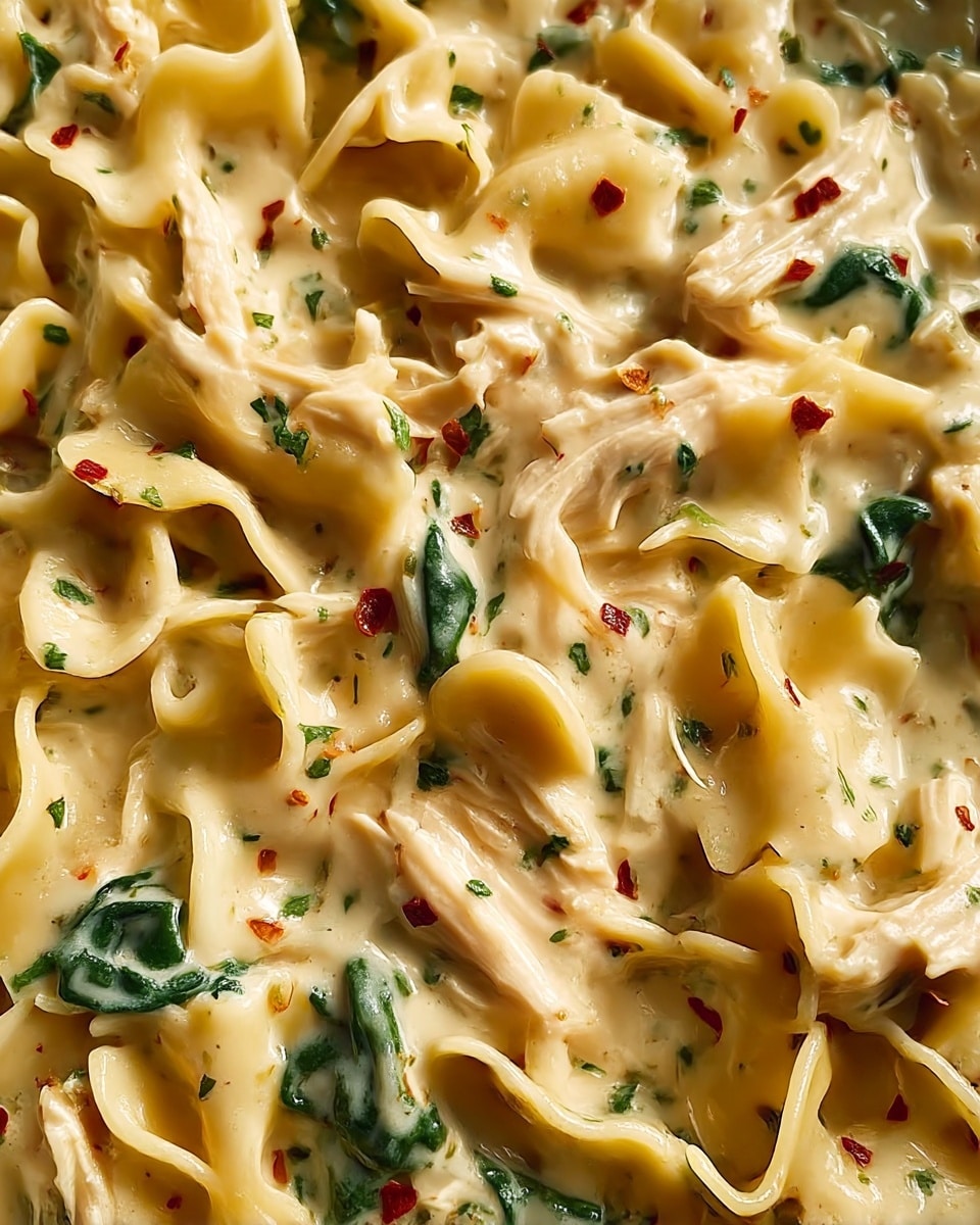 This close-up image shows creamy pasta with ruffled egg noodles mixed with shredded white chicken pieces and wilted dark green spinach throughout. The sauce is thick, smooth, and light beige, coating every part of the pasta and chicken evenly, with small bits of green herbs and red chili flakes sprinkled on top for a touch of color and texture. The overall look is rich and comforting, with the noodles curling gently and layering naturally within the creamy sauce. photo taken with an iphone --ar 4:5 --v 7