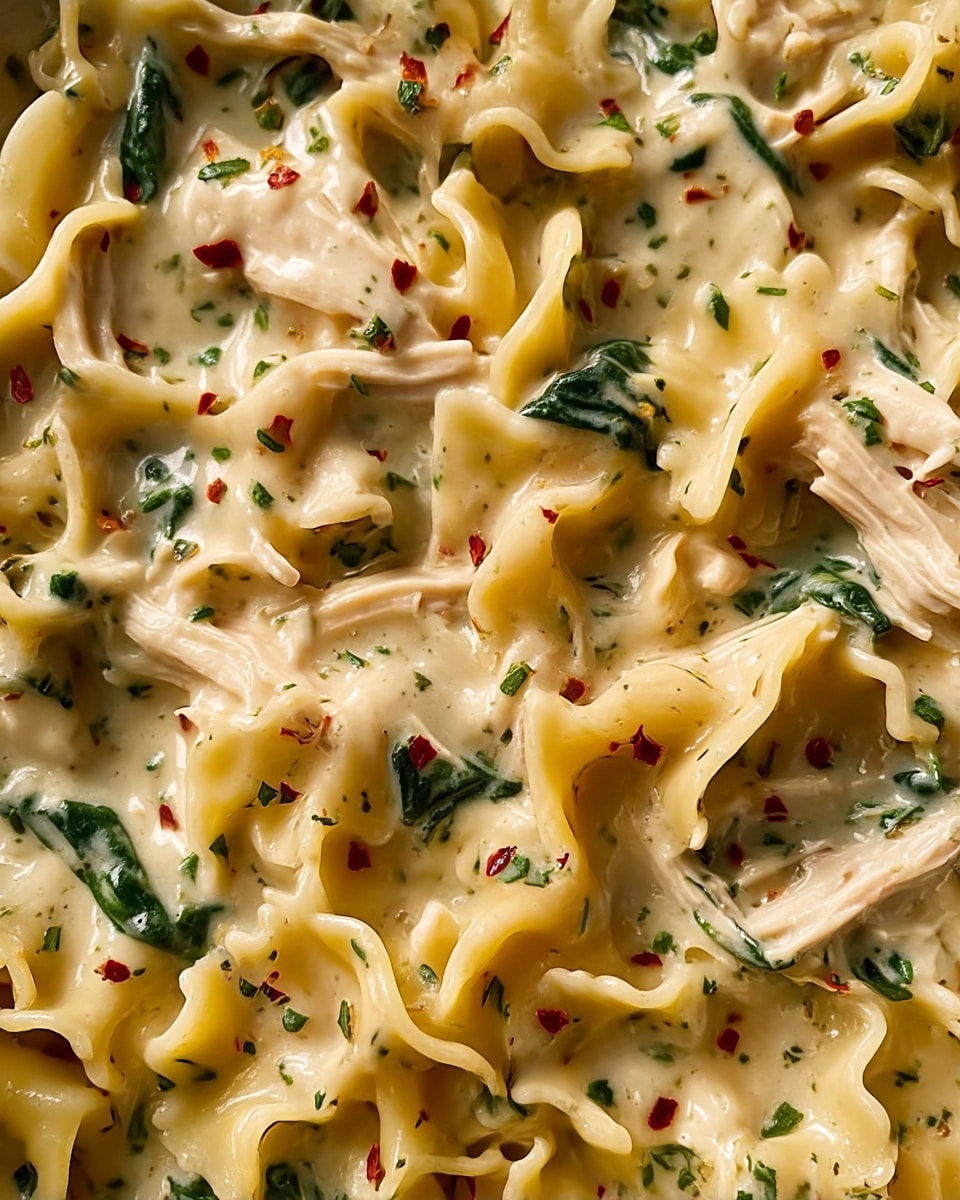 A close-up view of creamy pasta with three main layers: the bottom layer has curled yellow pasta ribbons with soft edges, the middle layer is a rich, thick white sauce dotted with finely chopped dark green herbs and small pieces of red flakes, and the top layer includes tender white chicken pieces and scattered leafy spinach adding dark green pops. The sauce looks smooth, glossy, and mixed evenly among the pasta and chicken. The image shows a fresh, warm texture with herbs and chili flakes spread lightly over the whole dish. Photo taken with an iphone --ar 4:5 --v 7