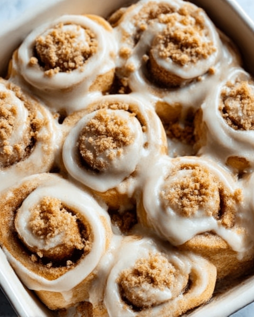A white baking dish filled with nine soft cinnamon rolls arranged in three rows, each roll swirled with light brown cinnamon filling. The rolls are covered in a thick, creamy, off-white glaze that pools slightly between them and glistens under the light. Crumbled bits of light brown streusel topping are scattered over the center of the cinnamon rolls, adding a crumbly texture contrast. The dish is set on a white marbled surface. photo taken with an iphone --ar 4:5 --v 7